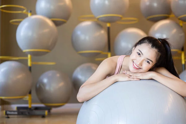 mulher debruçada em bola de pilates sorrindo para a camera